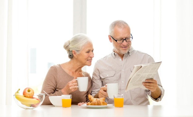 Concetto di famiglia, tecnologia, cibo, bevande e persone - coppia senior felice che fa colazione e legge il giornale a casa