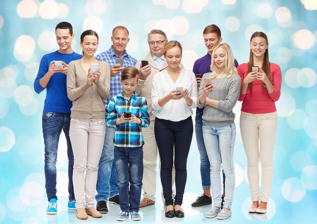 family, technology, generation and people concept - group of smiling men, women and boy with smartphones over blue holidays lights background