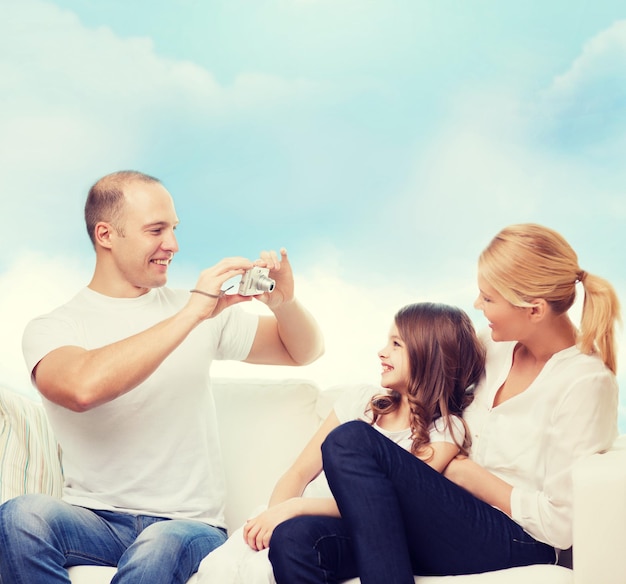 family, technology and people - smiling mother, father and little girl with camera over blue sky background