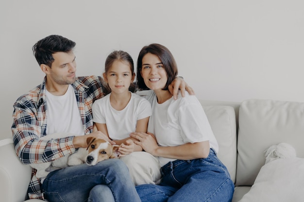 Family time concept cheerful mum dad and small girl embrace all
together spend spare time in living room of their new home sit on
sofa play with dog have perfect relationships enjoy comfort