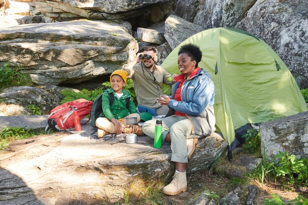 Famiglia di turisti che si godono la natura selvaggia e bevono tè seduti vicino alla tenda all'aperto si accampano