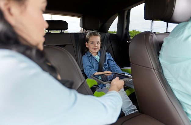 family, transport, road trip, travel and people concept - happy woman giving tablet pc computer to little daughter in car