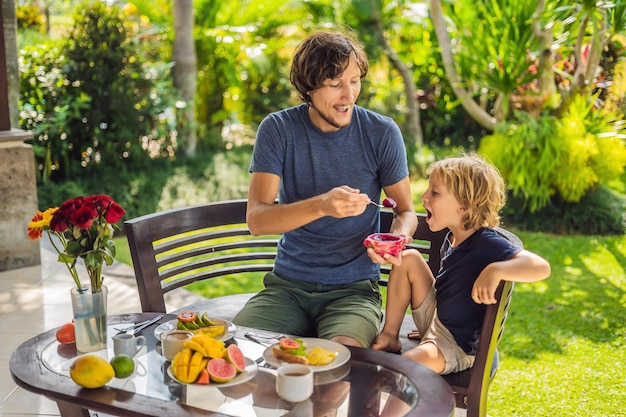 family of two eating nicely served breakfast outside handsome young man pouring some coffee and his cute son eating delicious fruit at breakfast time.