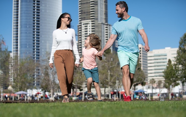 Family in urban skyline on park lawn Happy parents holding hands with son and walking in sunny summer citi street funny family moment