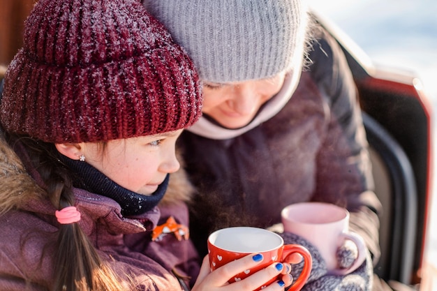 Family vacation family values Mother spends fun with her daughter in the winter in the park Mom and daughter are drinking tea while sitting in the open trunk of their SUV