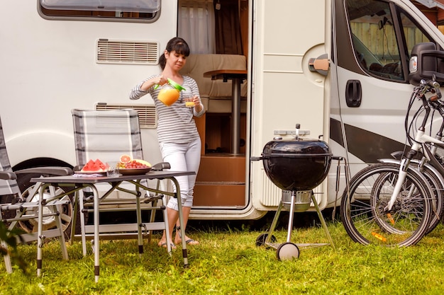 Family vacation travel RV, holiday trip in motorhome, Caravan car Vacation. Picnic with outdoor barbecue. Woman pours a glass of orange juice