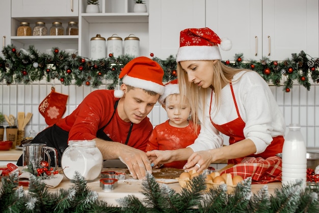 Family wearing santa hats cooking homemade cakes mom and dad are learning their son to cut out shapes from the dough