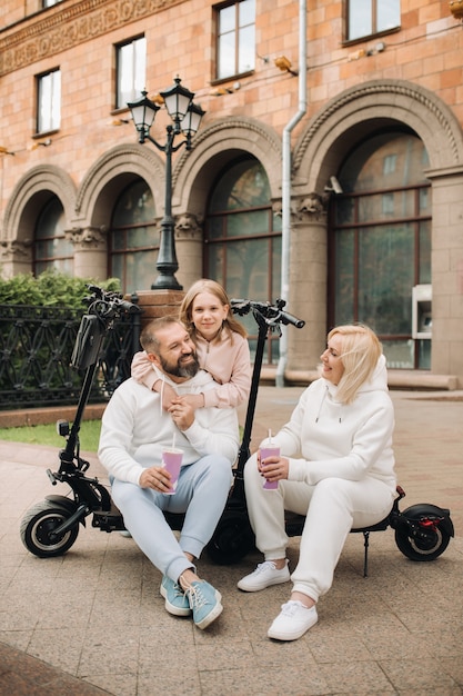 A family in white clothes stands in the city on electric scooters and drinks a drink.