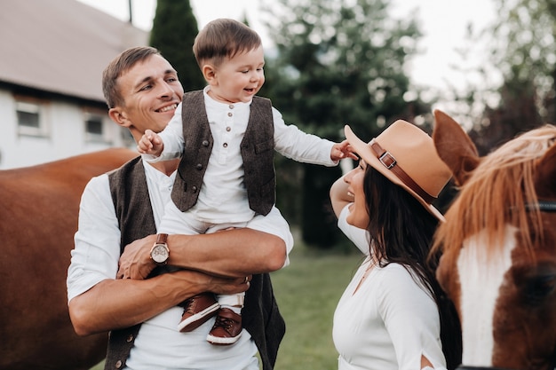 A family in white clothes with their son stand near two beautiful horses in nature. A stylish couple with a child are photographed with horses.