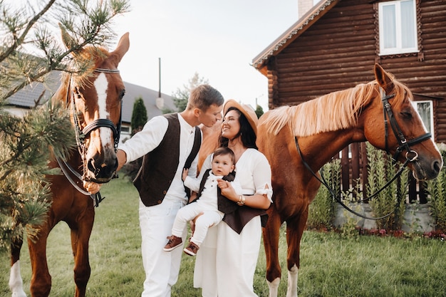 A family in white clothes with their son stand near two beautiful horses in nature. A stylish couple with a child are photographed with horses