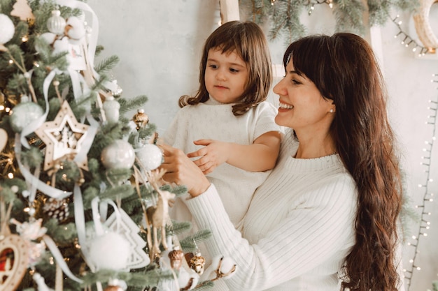Концепция семьи, зимних праздников и людей - счастливая мать держит маленькую дочь на руках и украшает елку дома