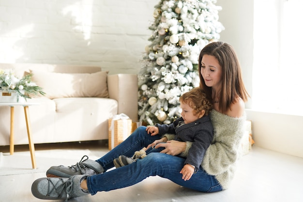 Семья, зимние каникулы и люди концепции - счастливая мама и малыш возле елки дома
