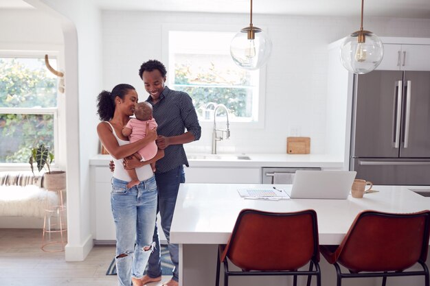 Family With Baby Daughter In Kitchen Using Laptop On Counter