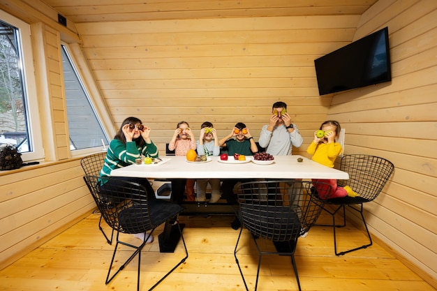 Family with four kids eat fruits in wooden country house on weekend They put fruit to their eyes