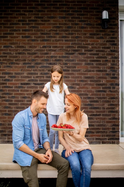 Photo family with a mother father and daughter sitting outside on a steps of a front porch of a brick house and eating strawberries
