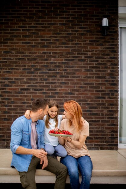 Family with a mother father and daughter sitting outside on a steps of a front porch of a brick house and eating strawberries