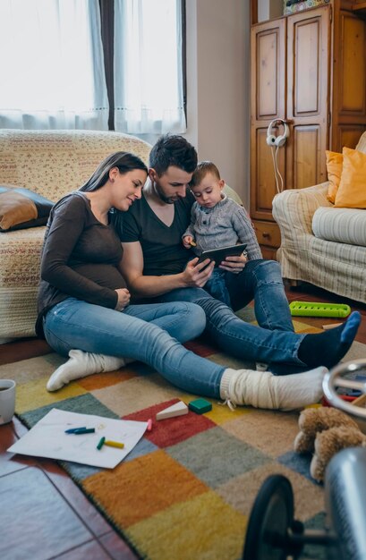 Family with small child and pregnant mother looking at the tablet sitting on the carpet