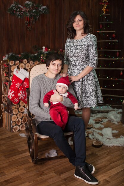 Una famiglia con un bambino piccolo in costume da babbo natale a casa davanti a un caminetto su una sedia a dondolo