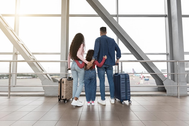 Family with suitcases looking out window at airport while waiting for flight