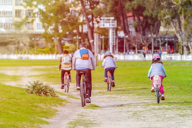 写真 安全な距離、背面図、暖かい色で調子を保つ都市公園で自転車に乗る2人の娘を持つ家族