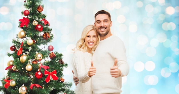 family, x-mas, winter holidays and people concept - happy couple showing thumbs up with christmas tree at home over blue lights background