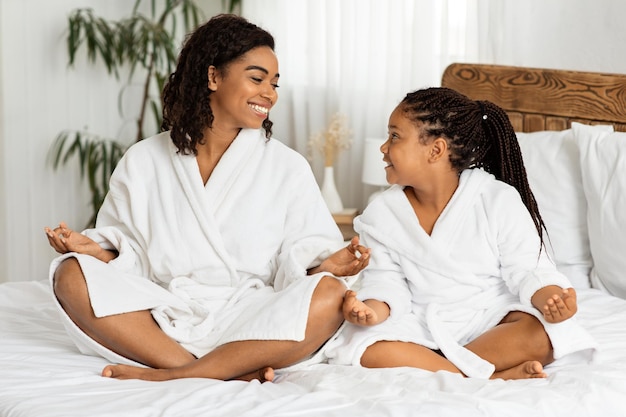 ファミリーヨガ。幸せな黒人のママと娘が自宅で一緒に瞑想するバスローブを着て、入浴後にロータスの位置でベッドに座って、お互いに絆を深め、笑顔で、寝室で休んで、空きスペース