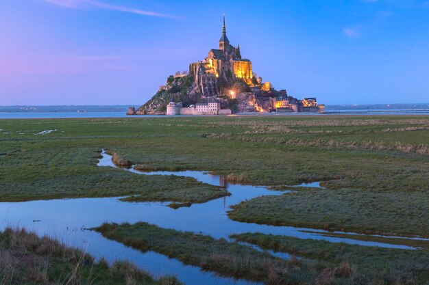 유명한 몽생미셸(Mont Saint Michel)은 프랑스 노르망디(Normandy)의 물 초원에 있는 운하에 반사되어 저녁 푸른 시간에 조명을 받았습니다.