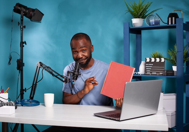 Famous vlogger reviewing excellent book talking to audience using professional microphone sitting at desk in vlogging studio. Content creator presenting a new novel volume to fanbase on social media.