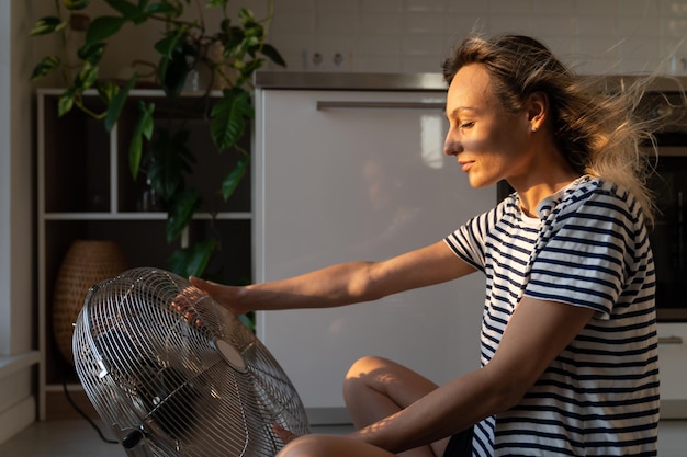 Fan blows female loose hair and cools down woman body sitting on floor in hot apartment in summer