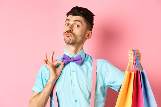 Ragazzo fantasia con i baffi che fissa il suo papillon e che tiene le borse della spesa, il ragazzo porta pacchetti regalo, in piedi su sfondo rosa.