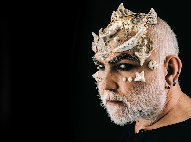 Fancy halloween makeup actor portraying ugly monster with\
thorns on face side view bearded man with evil eyes wearing silver\
ear rings isolated on black background