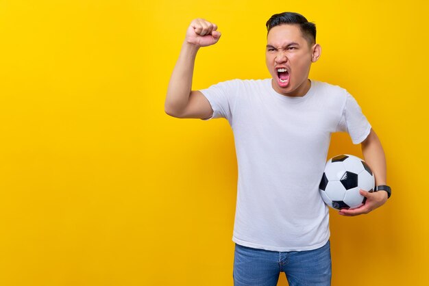 Fans excited to support the football sports team asian man 20s\
wears white tshirt holding a soccer ball and watching the live\
stream on tv with doing a winner gesture isolated on yellow\
background