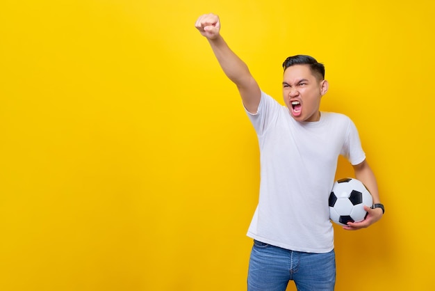 Fans excited to support the football sports team asian man 20s\
wears white tshirt holding a soccer ball and watching the live\
stream on tv with doing a winner gesture isolated on yellow\
background