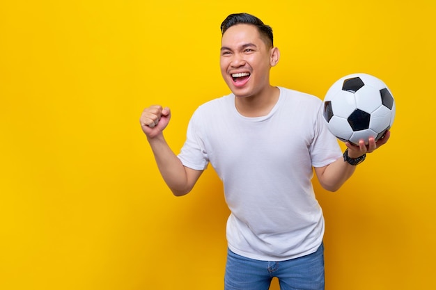 Fans excited to support the football sports team asian man 20s\
wears white tshirt holding a soccer ball and watching the live\
stream on tv with doing a winner gesture isolated on yellow\
background