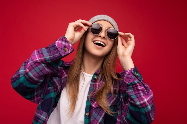 Fantastic closeup photo of amazing beautiful happy young dark blonde woman isolated over red background wearing colourful stylish shirt grey hat and modern sunglasses looking up and having fun