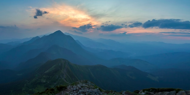 Fantastic sunrise or sunset over green mountain ridge covered with dense blue fog. Bright orange sun raising in  soft cloudy sky over distant horizon. Beauty of nature, tourism and traveling concept.