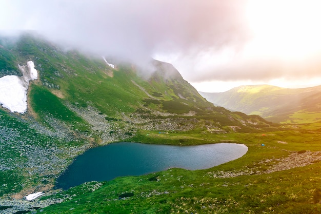 Fantastic wide peaceful view of blue lake in green valley and small tourist tents at rocky mountain with patches of snow covered with low clouds. Beauty of nature, tourism and traveling concept.