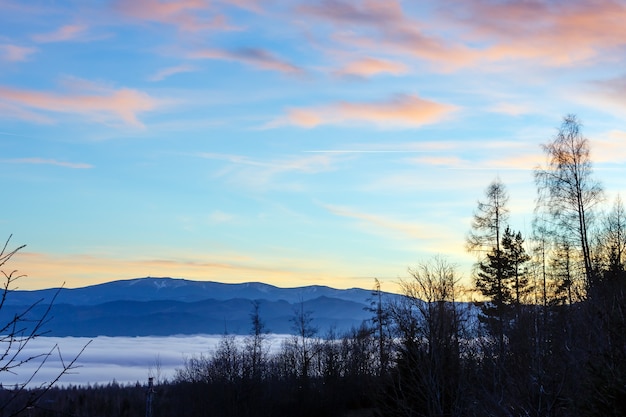 Fantastical sunset scenery with pink clouds illuminated by sun in sky and waves of clouds over foothills.