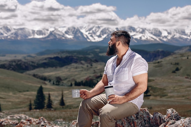 Fantastisch berglandschap en groene vallei. Een volwassen man met een baard in een wit overhemd zit op een geplaveide straat en houdt een mok en een thermoskan vast voor op reis. Kijk opzij naar het prachtige uitzicht.