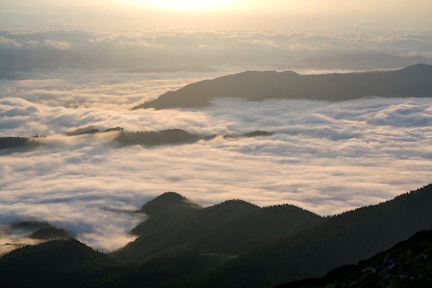 Fantastisch uitzicht op bergdal bedekt met lage witte gezwollen sneeuwwolken die zich uitstrekken tot mistige horizon onder heldere ochtendhemel met lichtoranje gloed bij zonsopgang. Schoonheid van de natuur concept.