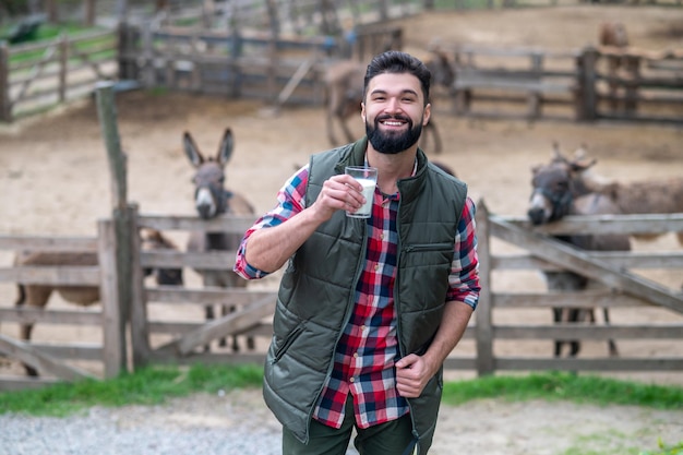 On a farm. A dark-haired bearded man in plaid shirt with a glass with milk standing near livestock