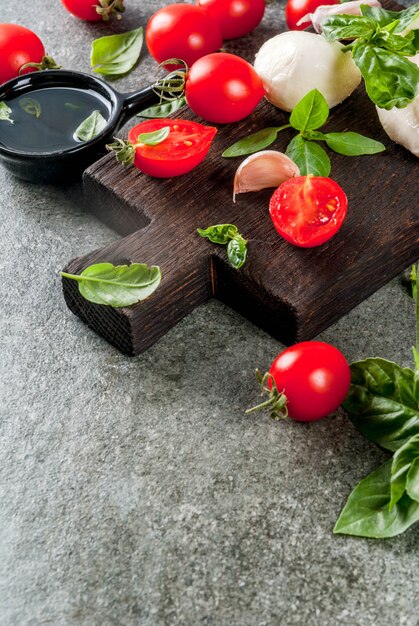 Farm rauwe biologische producten. Voorbereiding van het diner in de Italiaanse stijl. Ingrediënten voor Caprese Salade, Pasta, Pizza. Basilicum, tomaten, mozzarellakaas, olijfolie op donkere grijze stenen tafel. Kopieer ruimte