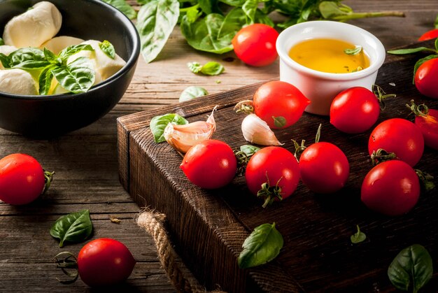 Farm raw organic products. Preparation of dinner in the Italian style. Ingredients for caprese salad, pasta, pizza. Basil, tomatoes, mozzarella cheese, olive oil on an old wooden table.