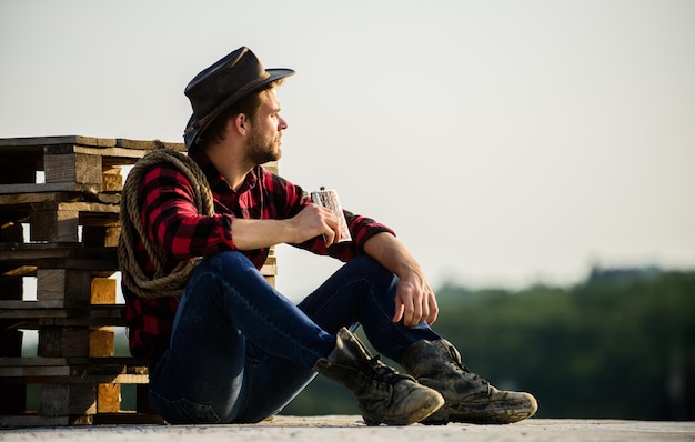 Farmer drink alcohol enjoy view from his farm Watching sunset Farmer cowboy handsome man relaxing after hard working day at ranch Romanticism western culture Farmer in hat sit relax Evening time