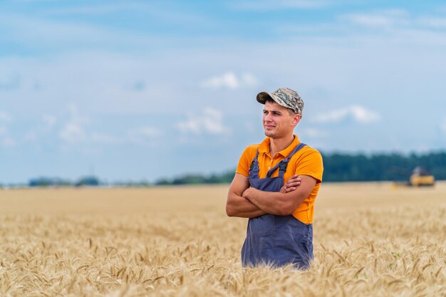 Farmer in orange and blue uniform standing in big beautiful yellow field with crossed arms and looking away. Breathtaking view of horizon and sky.