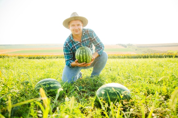 Фермер улыбается и смотрит в камеру, держа в руке большой арбуз, фермер в шляпе на ф ...