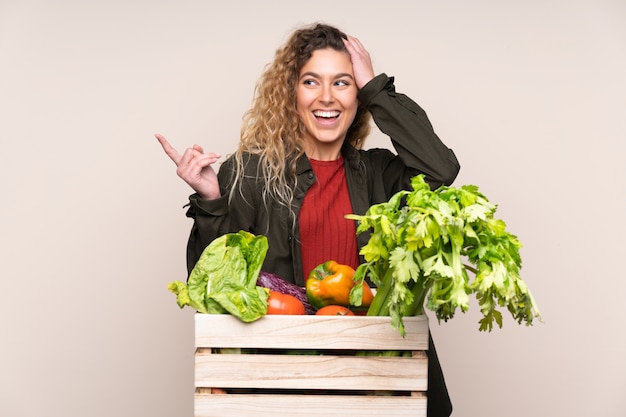 Farmer with freshly picked vegetables in a box on beige wall surprised and pointing finger to the side