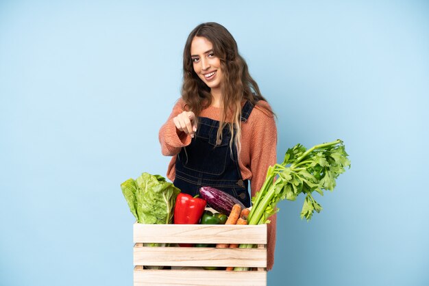 Farmer with freshly picked vegetables in a box points finger at you with a confident expression