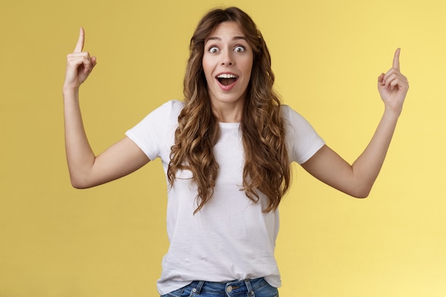 Photo fascinated speechless excited attractive curly-haired girl impressed stare camera surprised glad appreciation awesome gift receive incredible offer pointing up index fingers yellow background