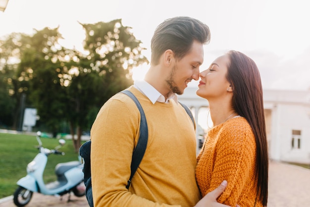 Fascinating girl with long dark hair spending weekend with boyfriend, walking in park. Charming white lady in orange sweater kissing bearded husband standing on blur background near scooter.